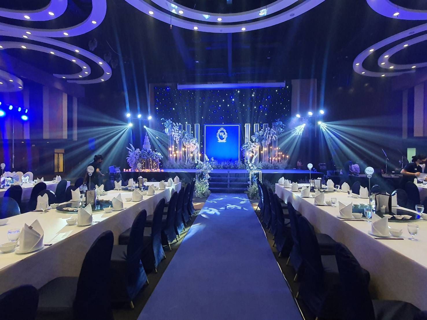 Banquet hall with tables set for a formal event; stage with blue lighting and decorations.