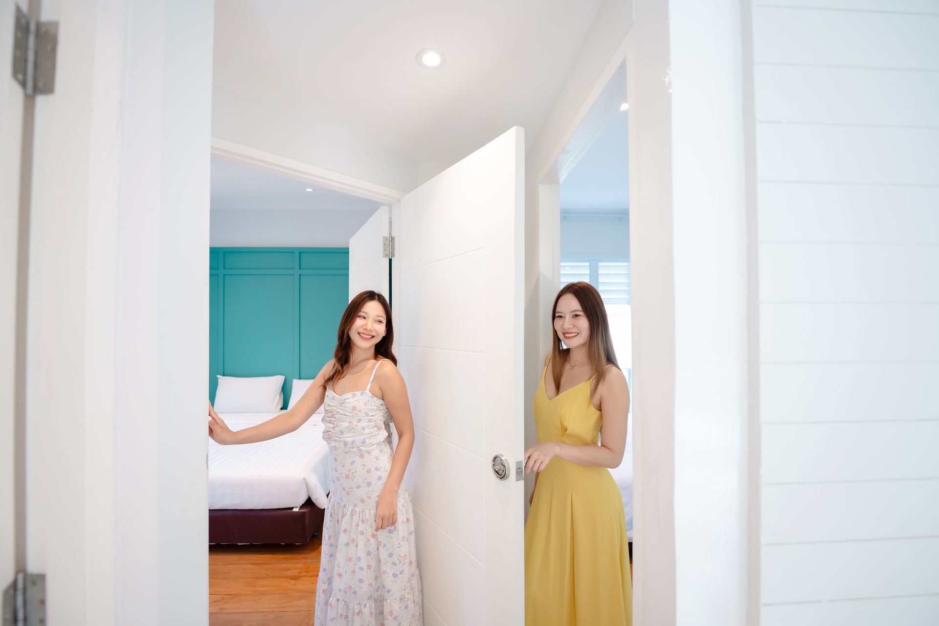 两名身穿连衣裙的女子打开一扇白色门，进入一间酒店房间，房间的墙壁和床都是蓝绿色的。