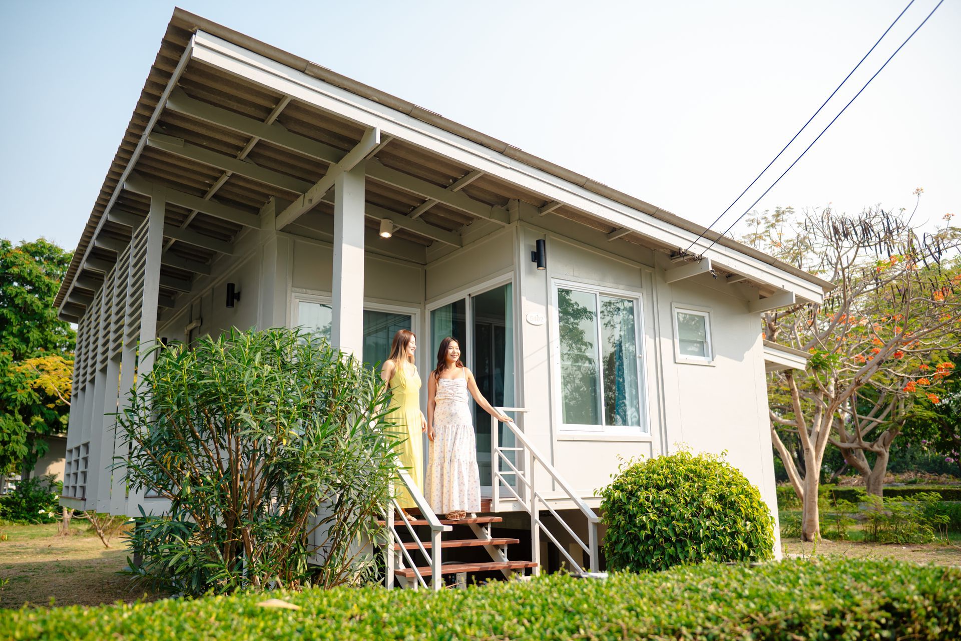 Two women stand on steps of a white house with a covered porch; green shrubs frame the house.