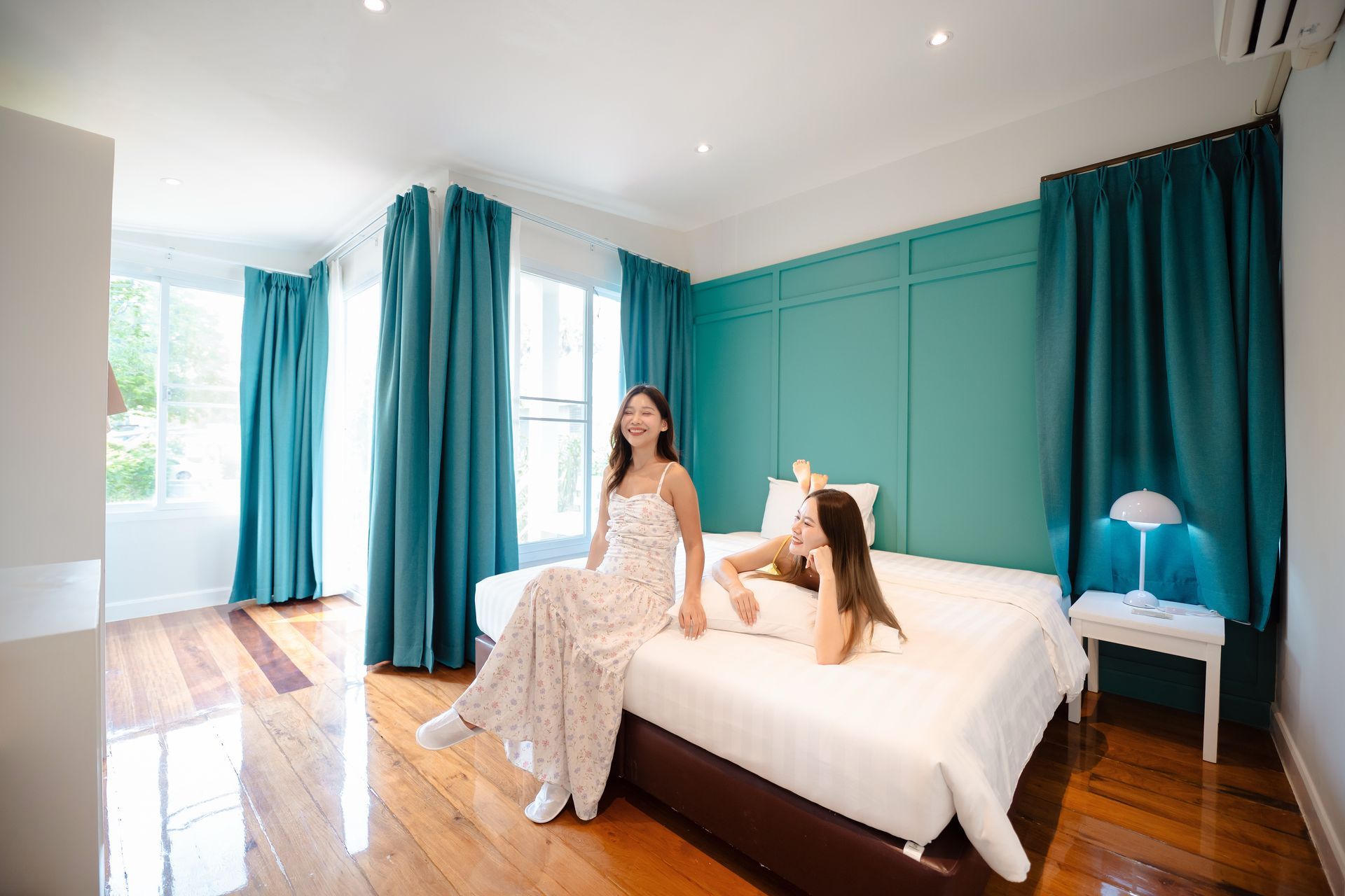 Two people on a bed in a teal-accented bedroom. One sits, the other reclines, both smiling.