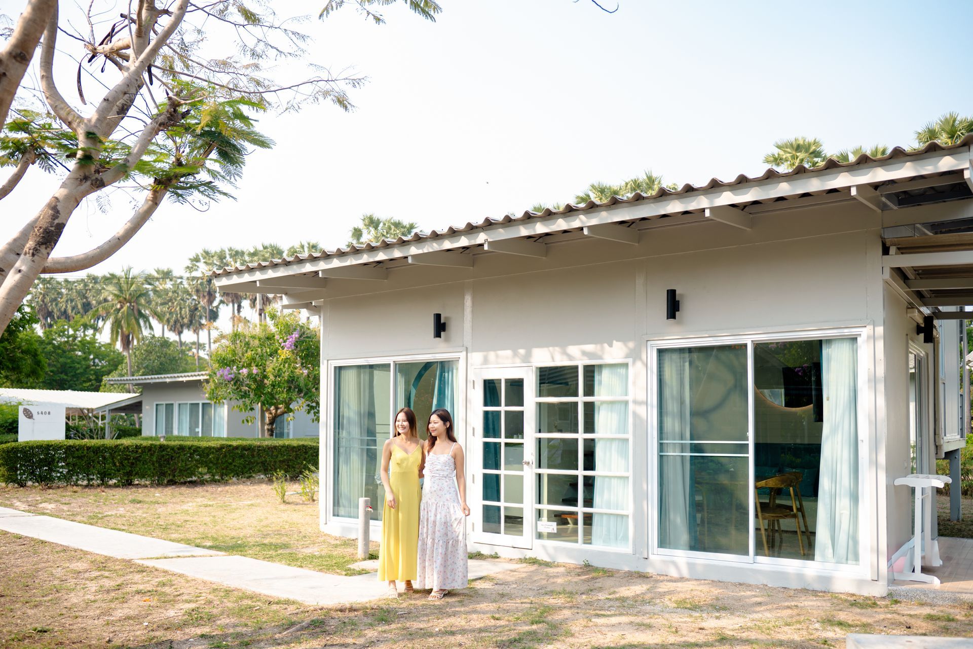 Two women in dresses walking in front of a white building with glass doors, sunny outdoors.