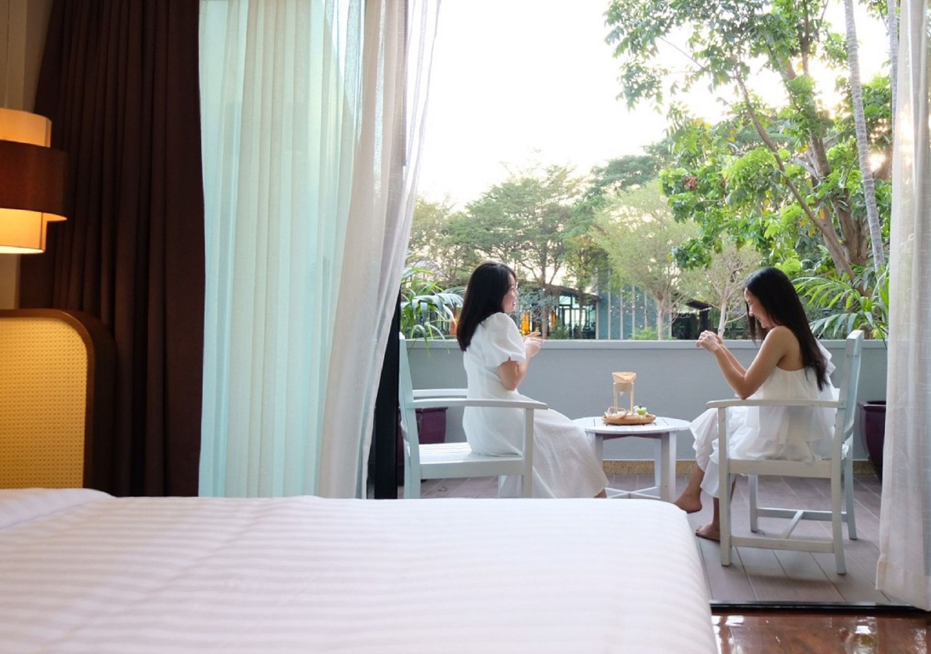 两位身穿白色连衣裙的女士在阳台上，一边啜饮饮品，一边欣赏绿意盎然的景色。