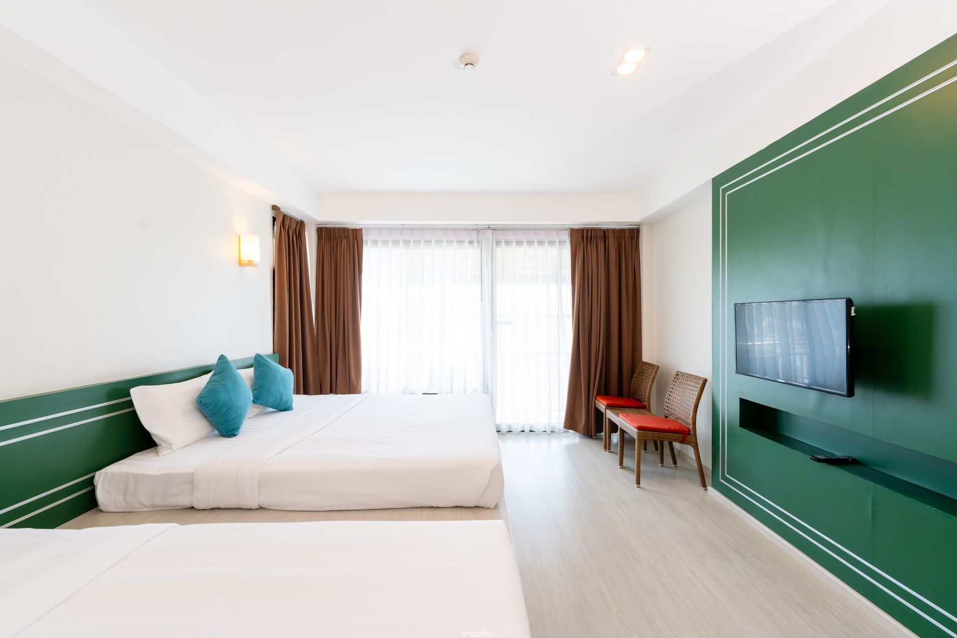 Hotel room with multiple beds, TV, and balcony access. Green wall accents, brown curtains, and light flooring.