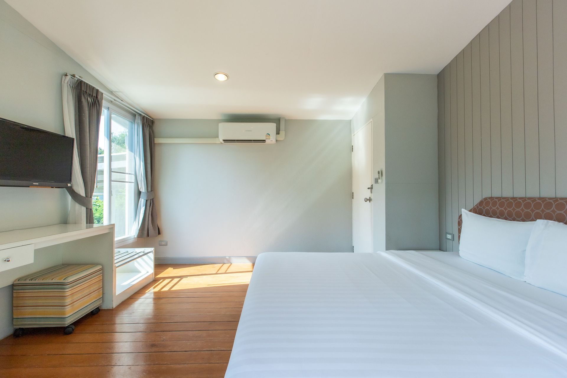 Bedroom with a bed, TV, desk, and window. Pale green walls, wooden floor, and white bedding.