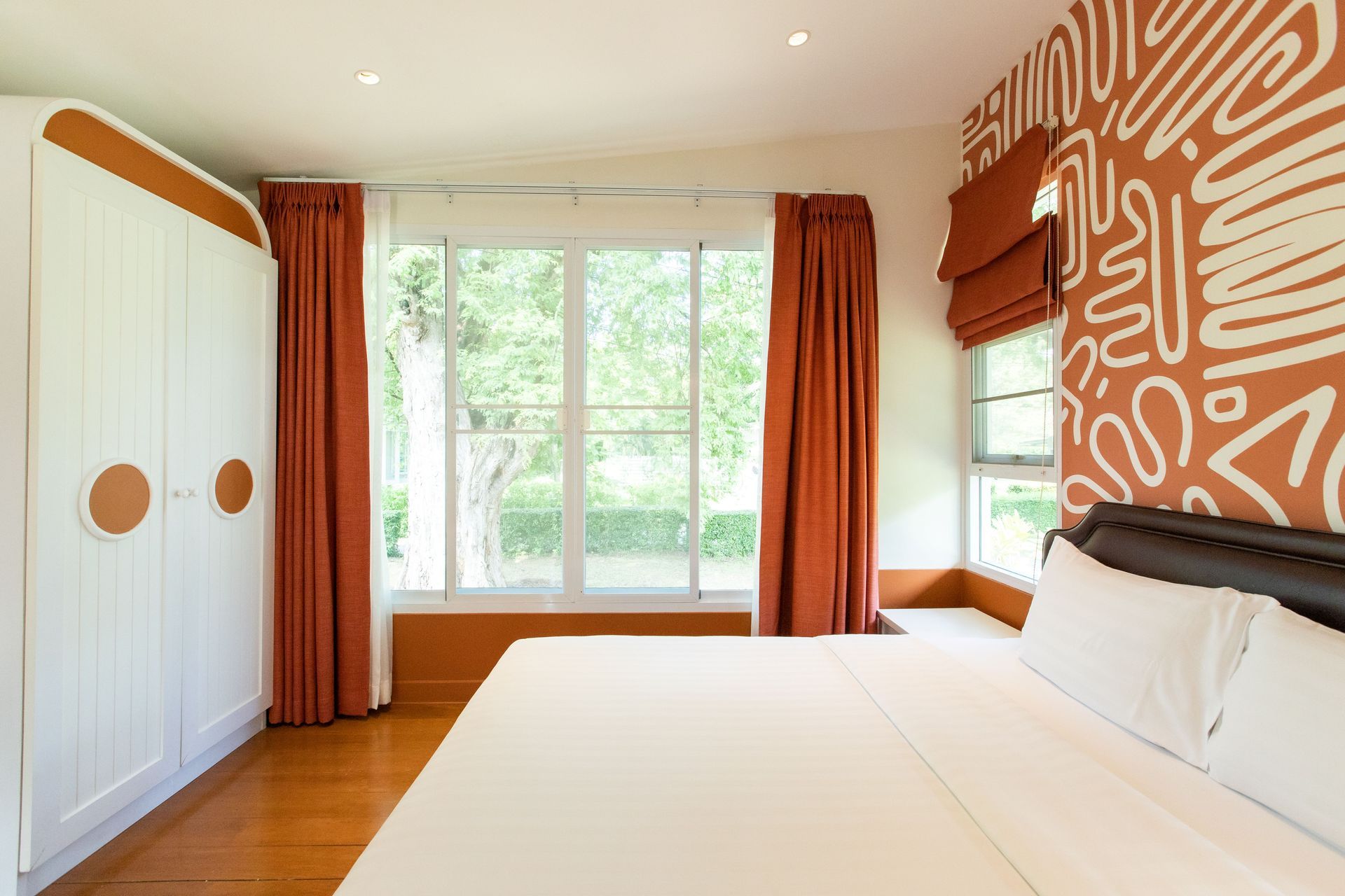 卧室里有一扇大窗户、一个白色衣柜和橙色窗帘。床上铺着白色床单。墙面是橙色的图案。