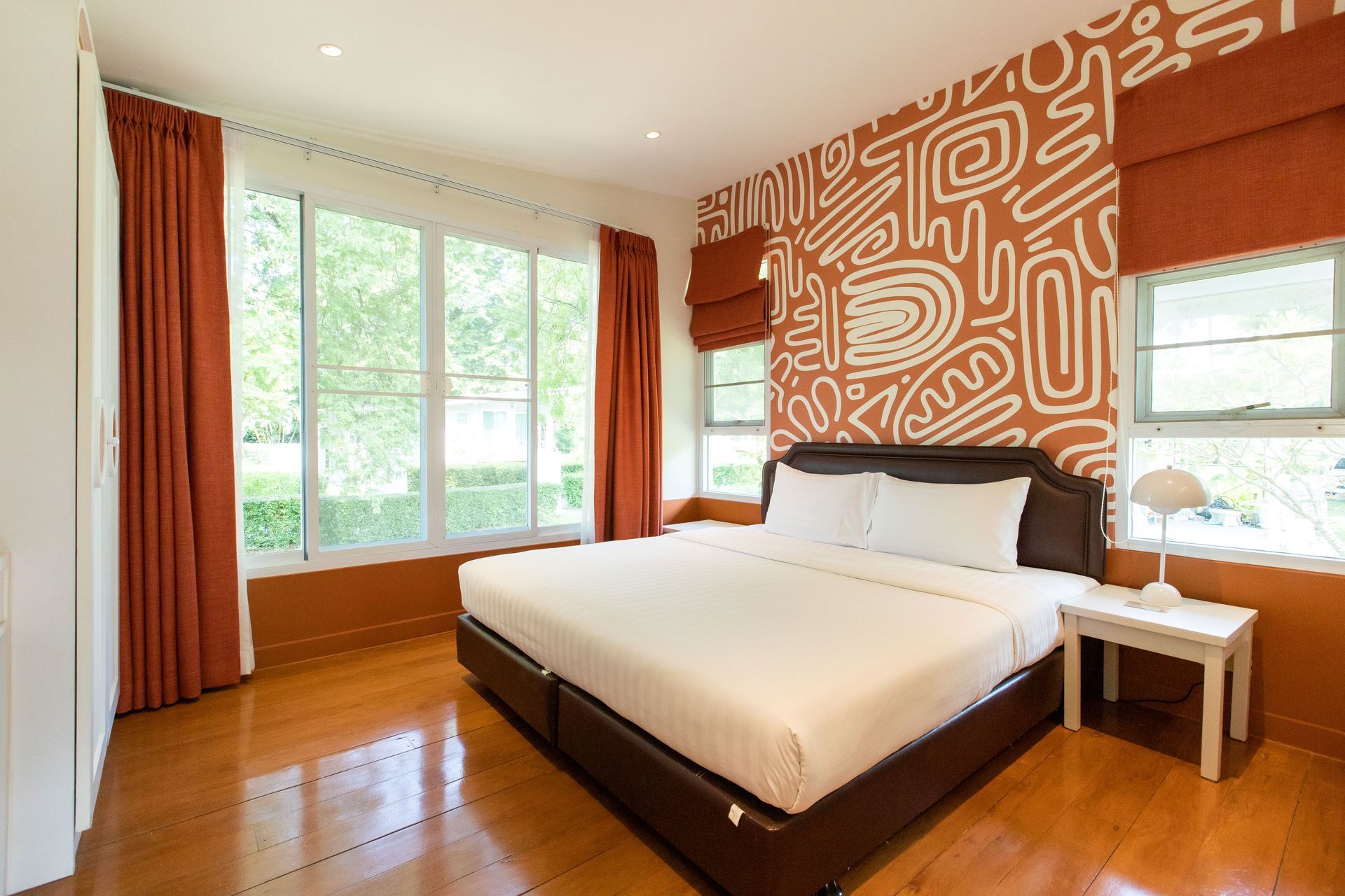 Bedroom with bed, patterned wall, windows, orange curtains, wooden floor.