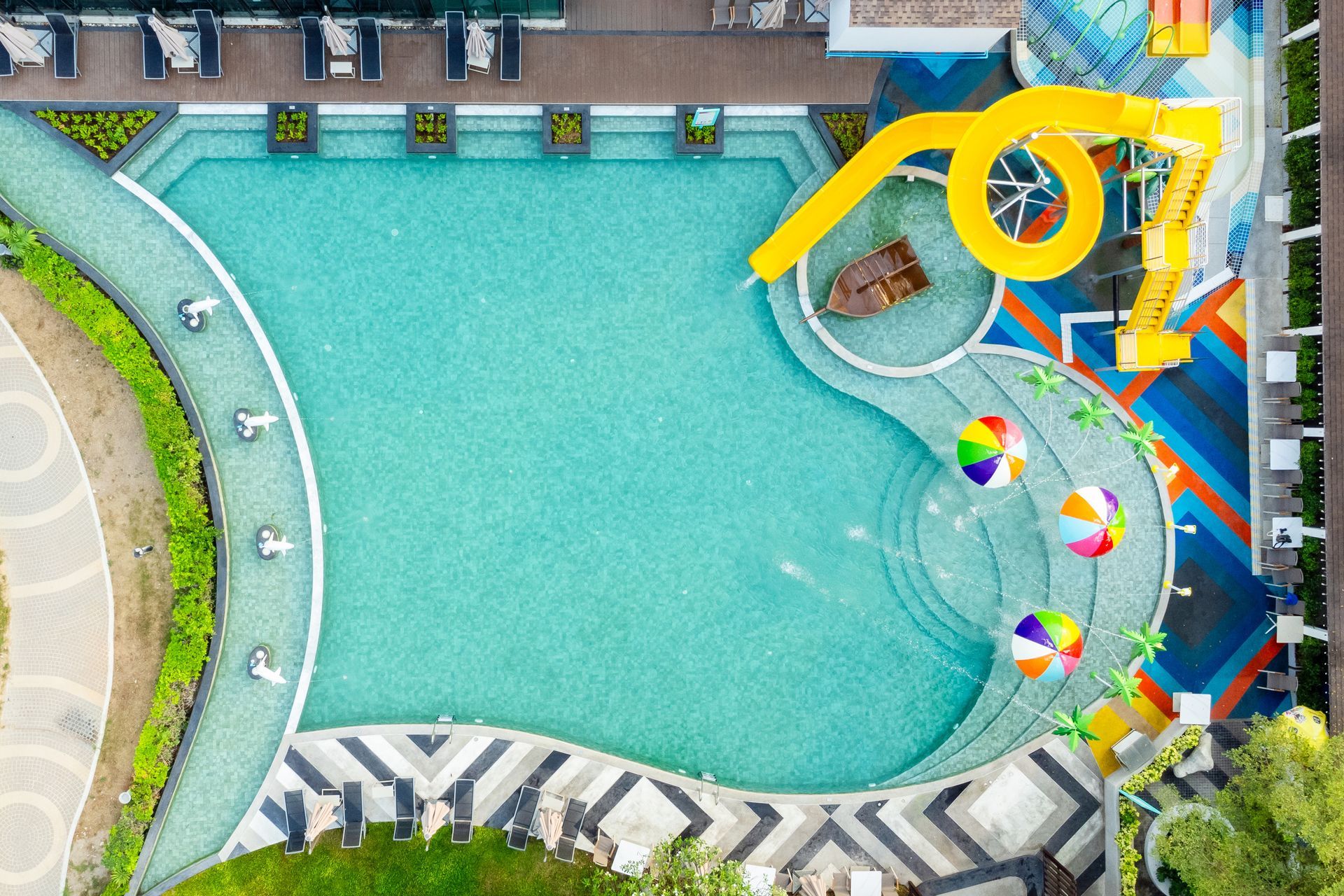 从空中俯瞰，可以看到一个游泳池，池边有黄色滑水道，周围环绕着色彩缤纷的遮阳伞和休闲区。