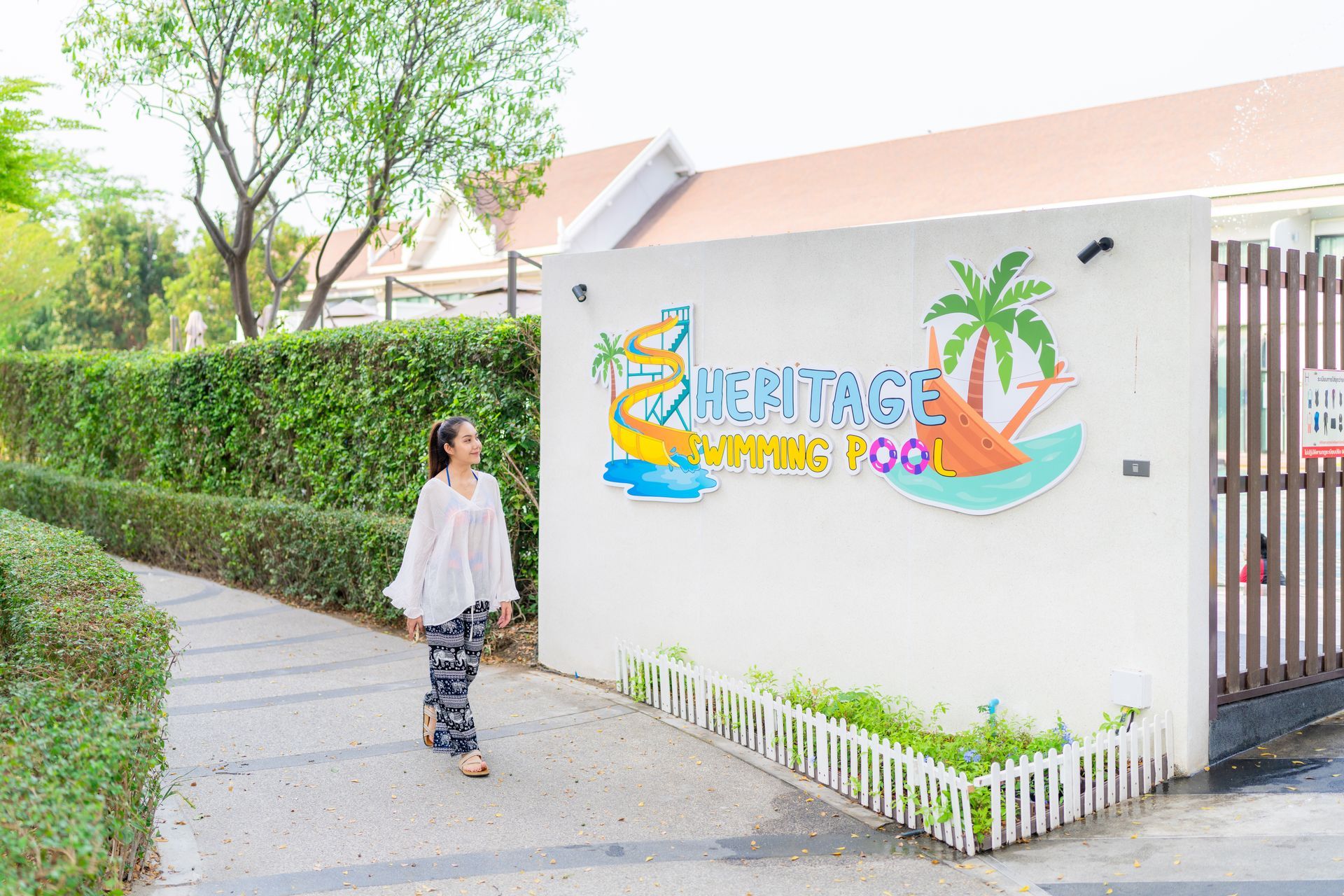 一位女士走过一块写着“遗产游泳池”的牌子，牌子上有大门、绿色树篱和树木。