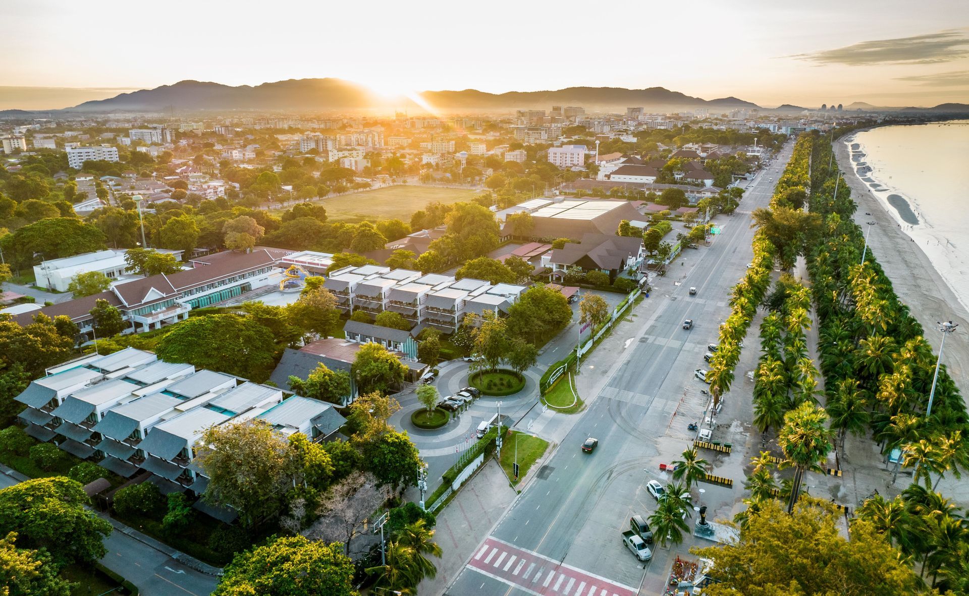 日出时分，从空中俯瞰一座沿海城市。一条道路将建筑物与海滩隔开，远处是连绵的群山。