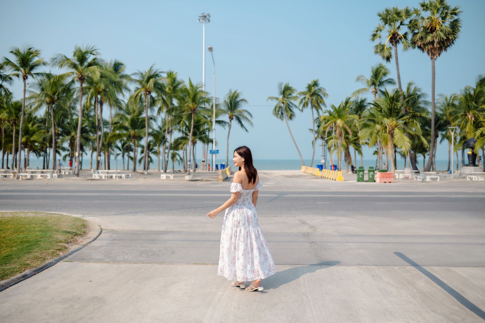 Woman in floral dress walking on a road towards a beach with palm trees on a sunny day.