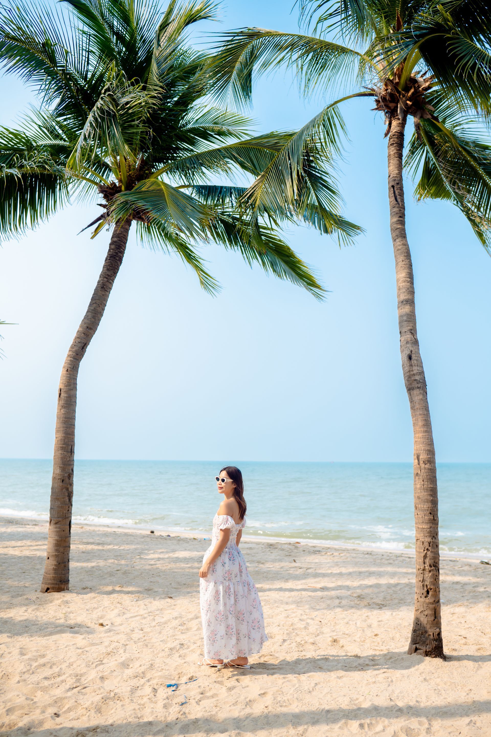 Woman in white dress stands on a sandy beach between two palm trees, ocean in the background, bright blue sky.