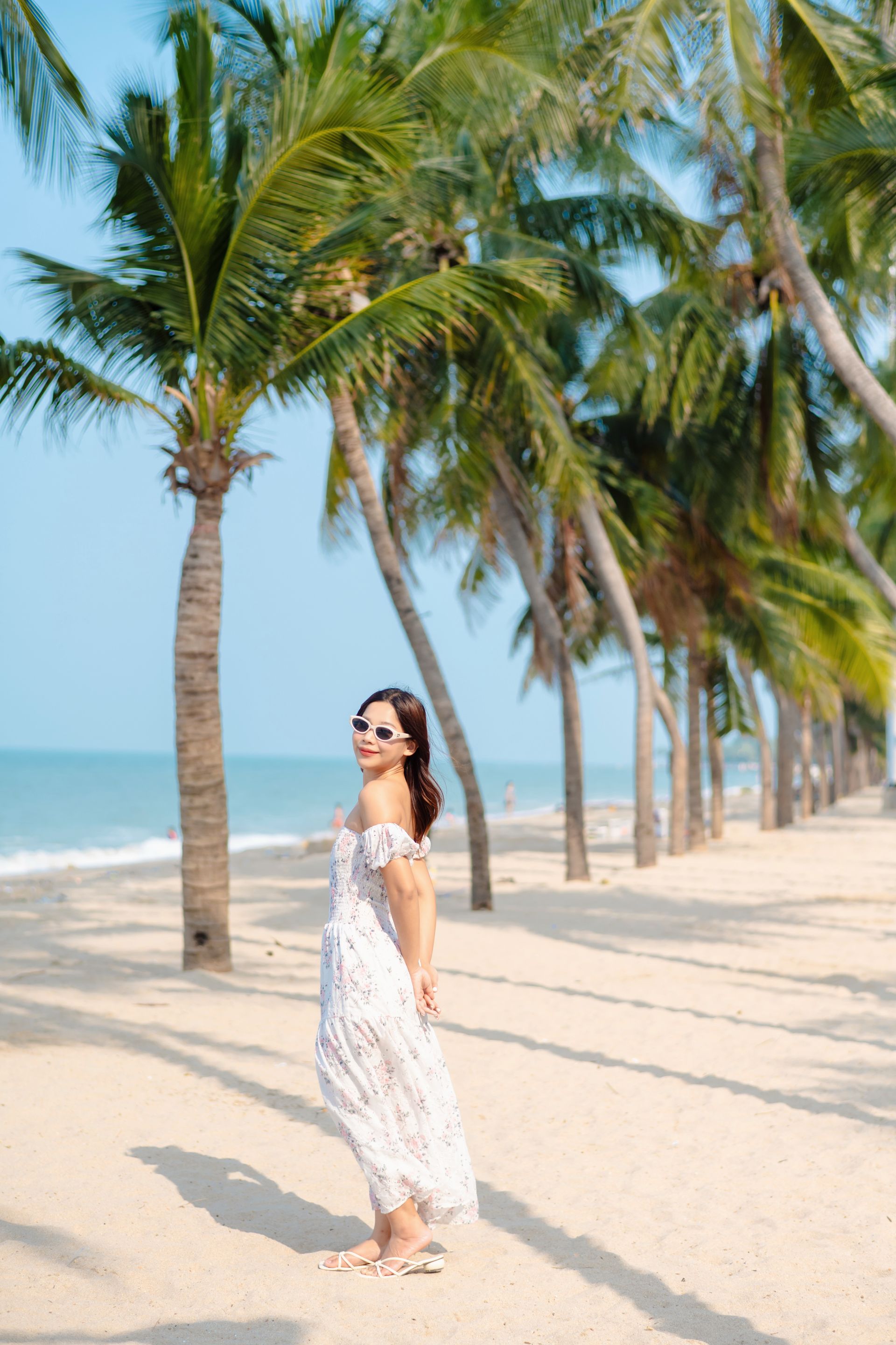 身穿白色连衣裙的女子面带微笑，站在棕榈树环绕的沙滩上。