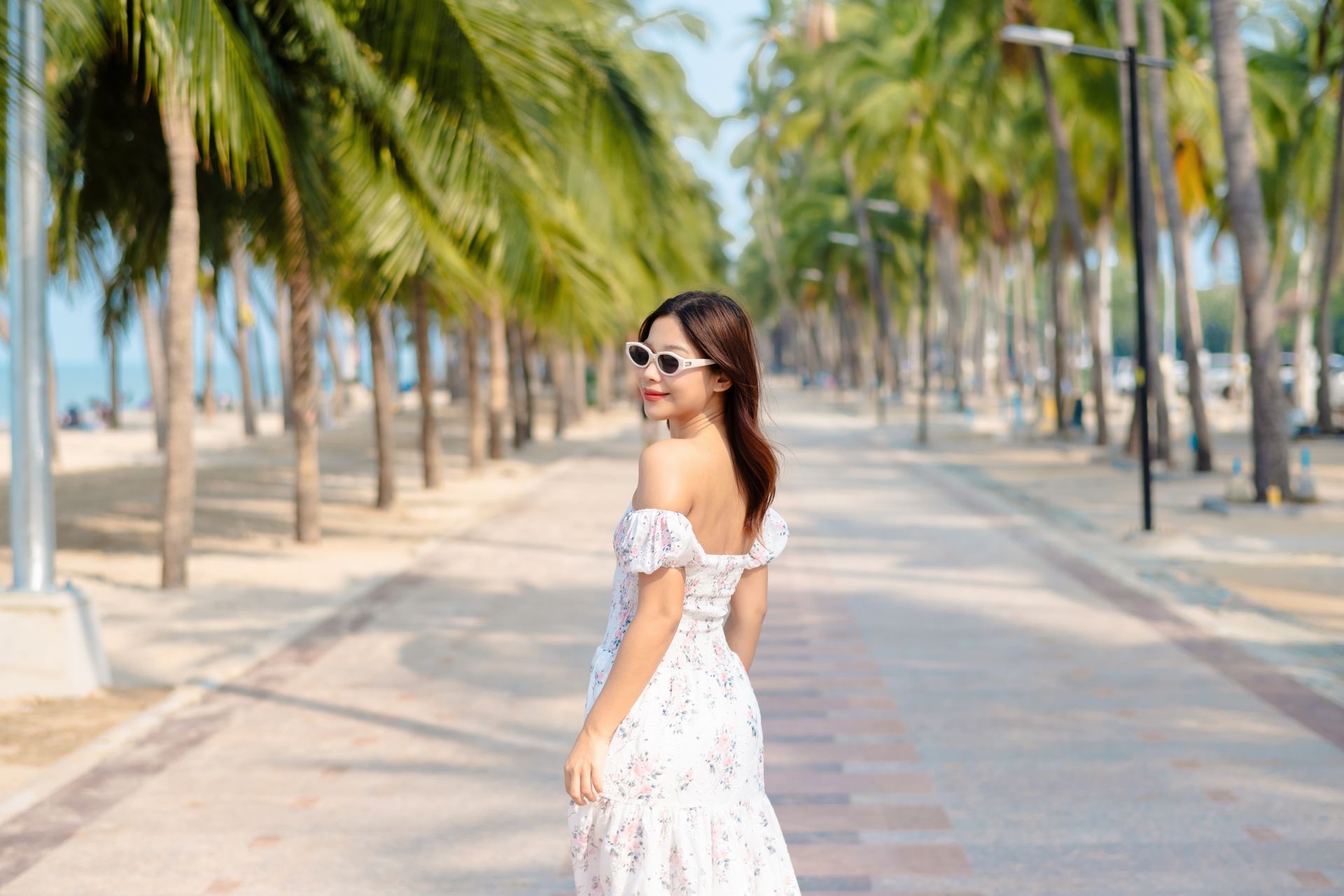 身穿白色连衣裙、戴着太阳镜的女子面带微笑，回头望向棕榈树荫下的小路。