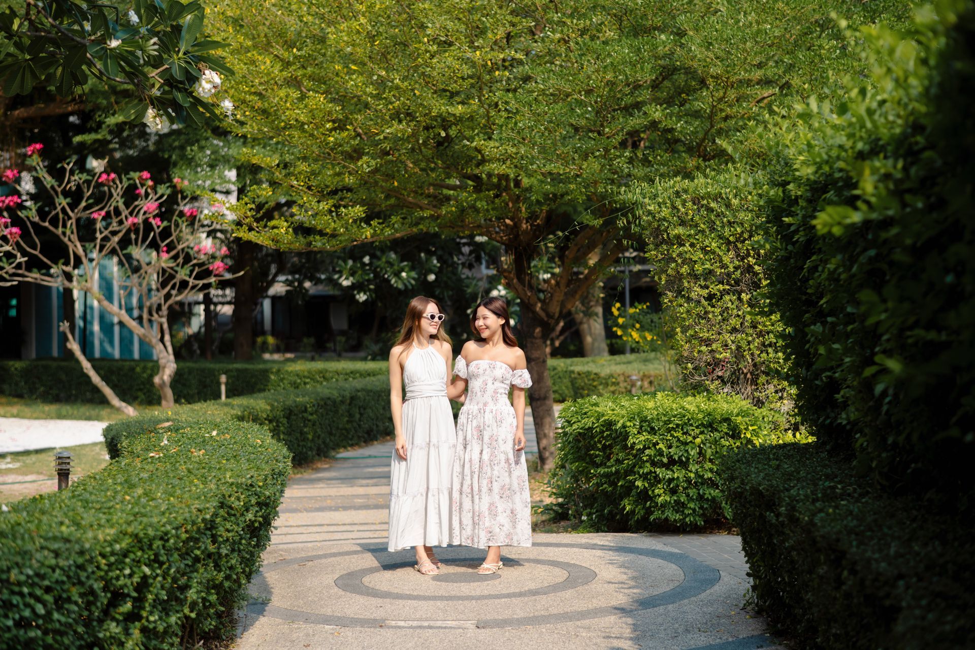 两名身穿白色连衣裙的女子走在花园的小路上，彼此微笑。