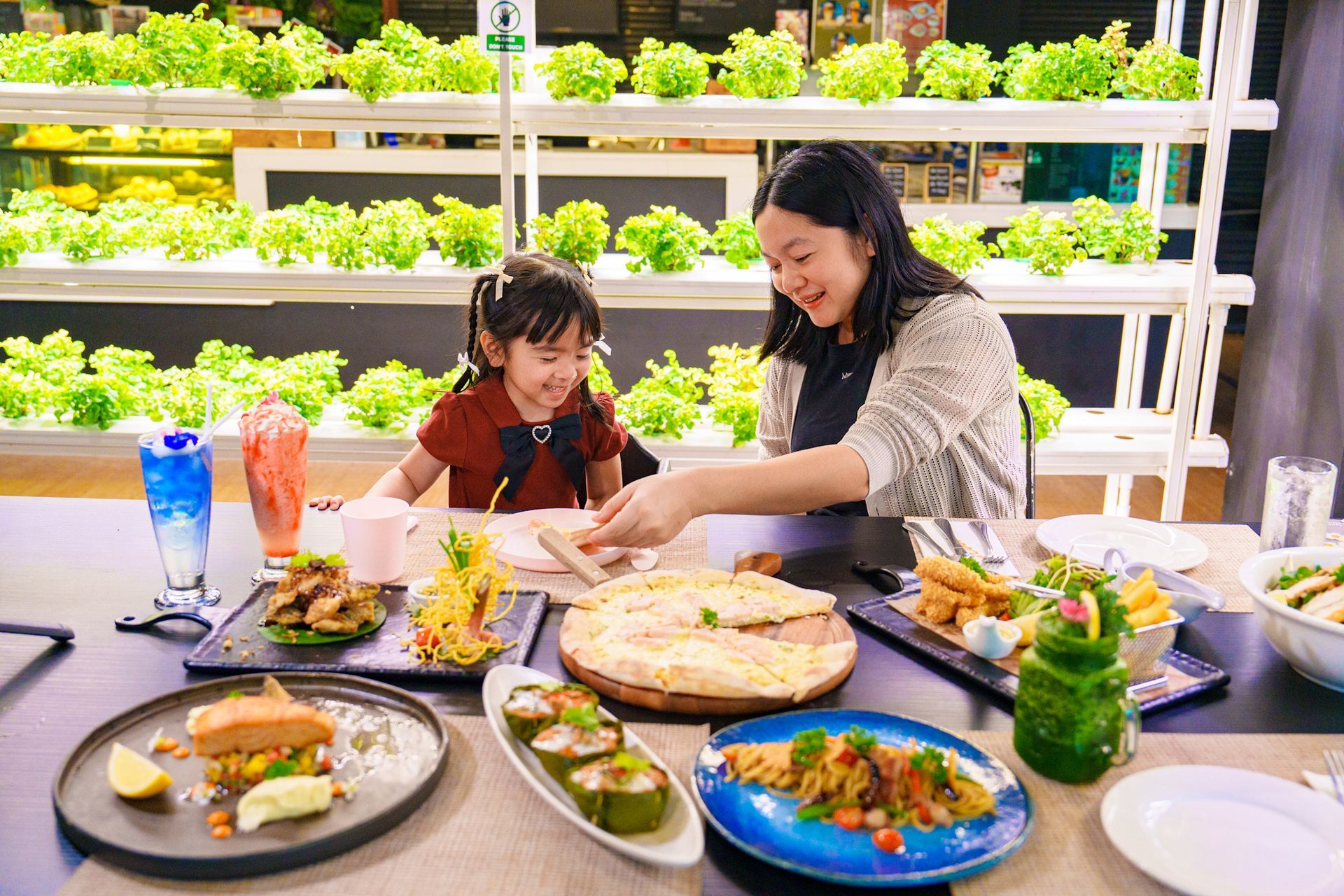 Woman and child eating at a table with various dishes in a restaurant with hydroponic plants in the background.