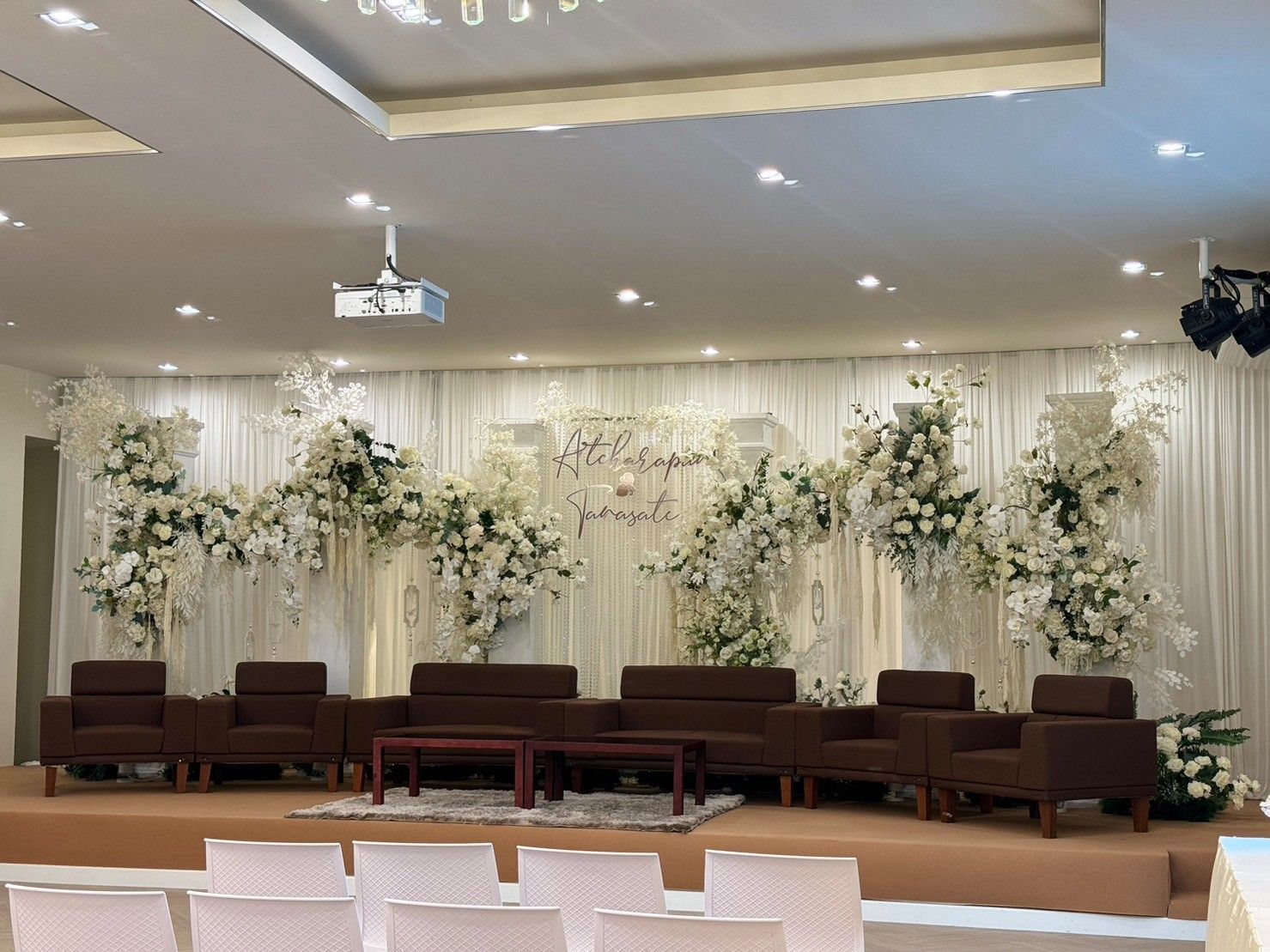 Stage decorated for an event, with white flowers, brown sofas, and white chairs in front.