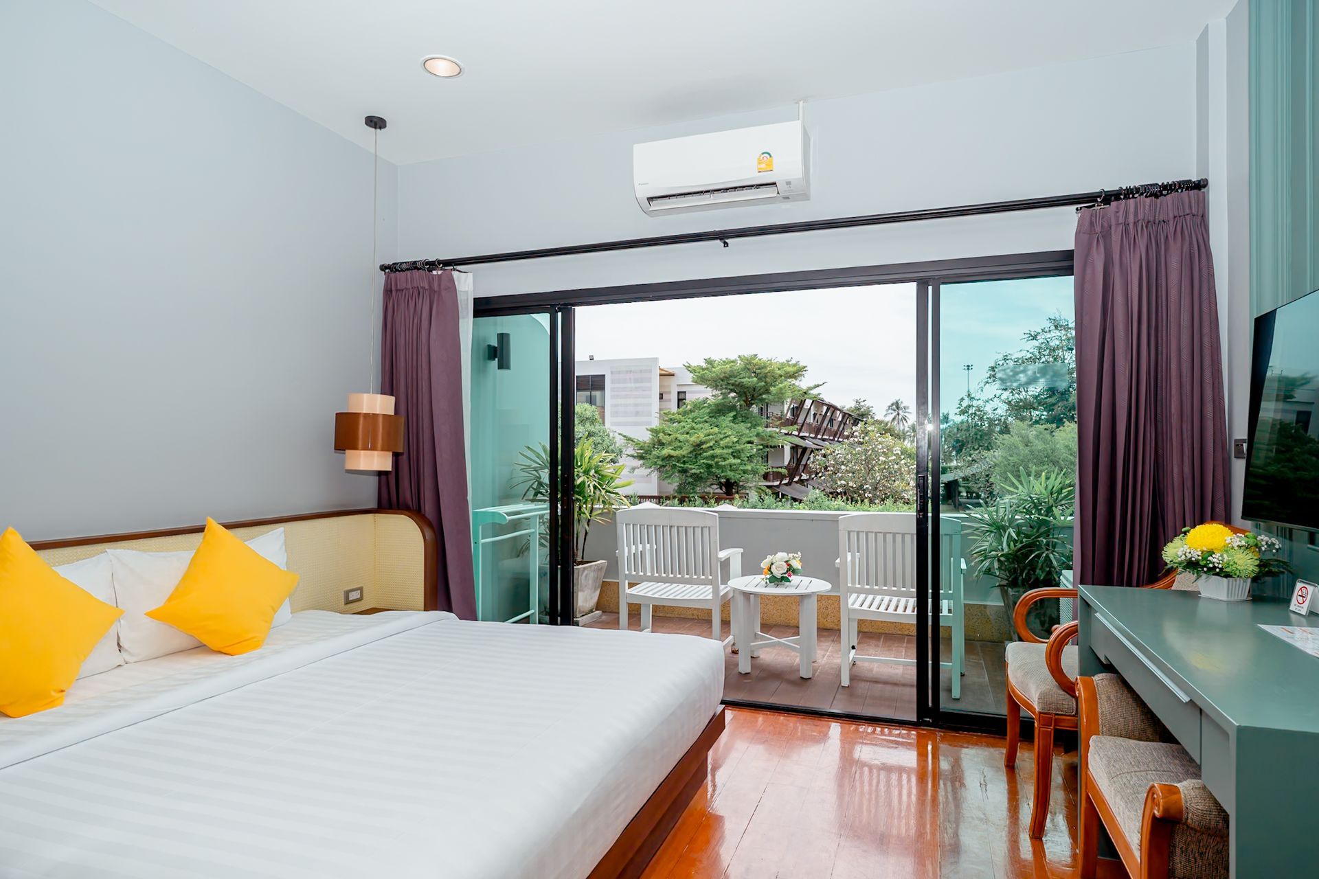 Bedroom with bed, balcony, and wooden floor. Bright, with yellow pillows and light purple curtains.