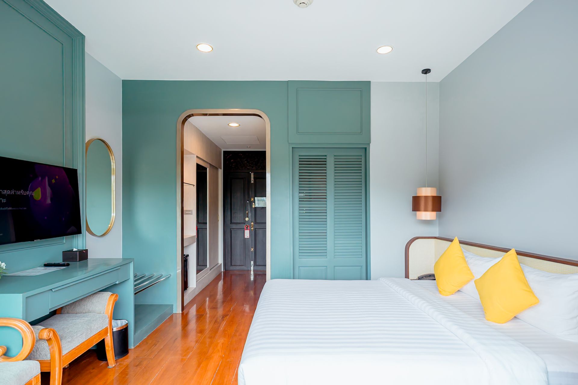 酒店房间，墙壁为蓝绿色和灰色，木地板，床上放着黄色枕头，桌子上有一台电视。