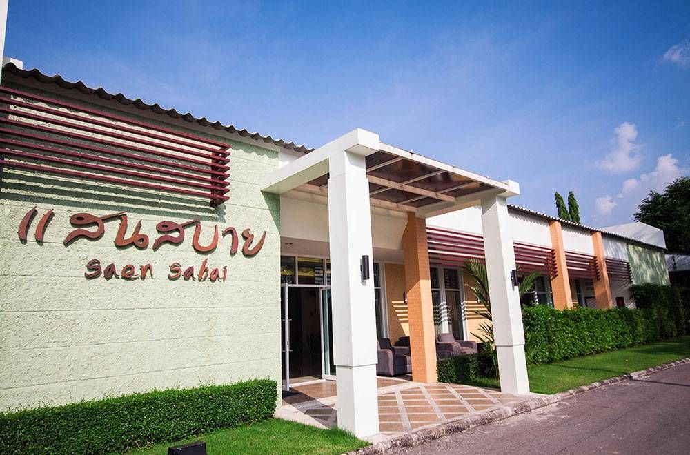 Saan Sabai 大楼入口处以棕色和白色为主色调，周围环绕着绿色的灌木丛和蓝色的天空。