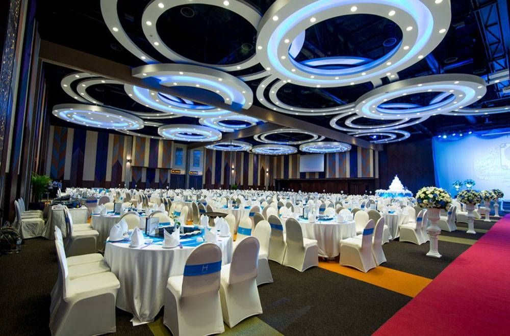 宴会厅布置妥当，准备举办活动；白色桌椅，蓝色点缀，装饰性天花板灯，舞台。