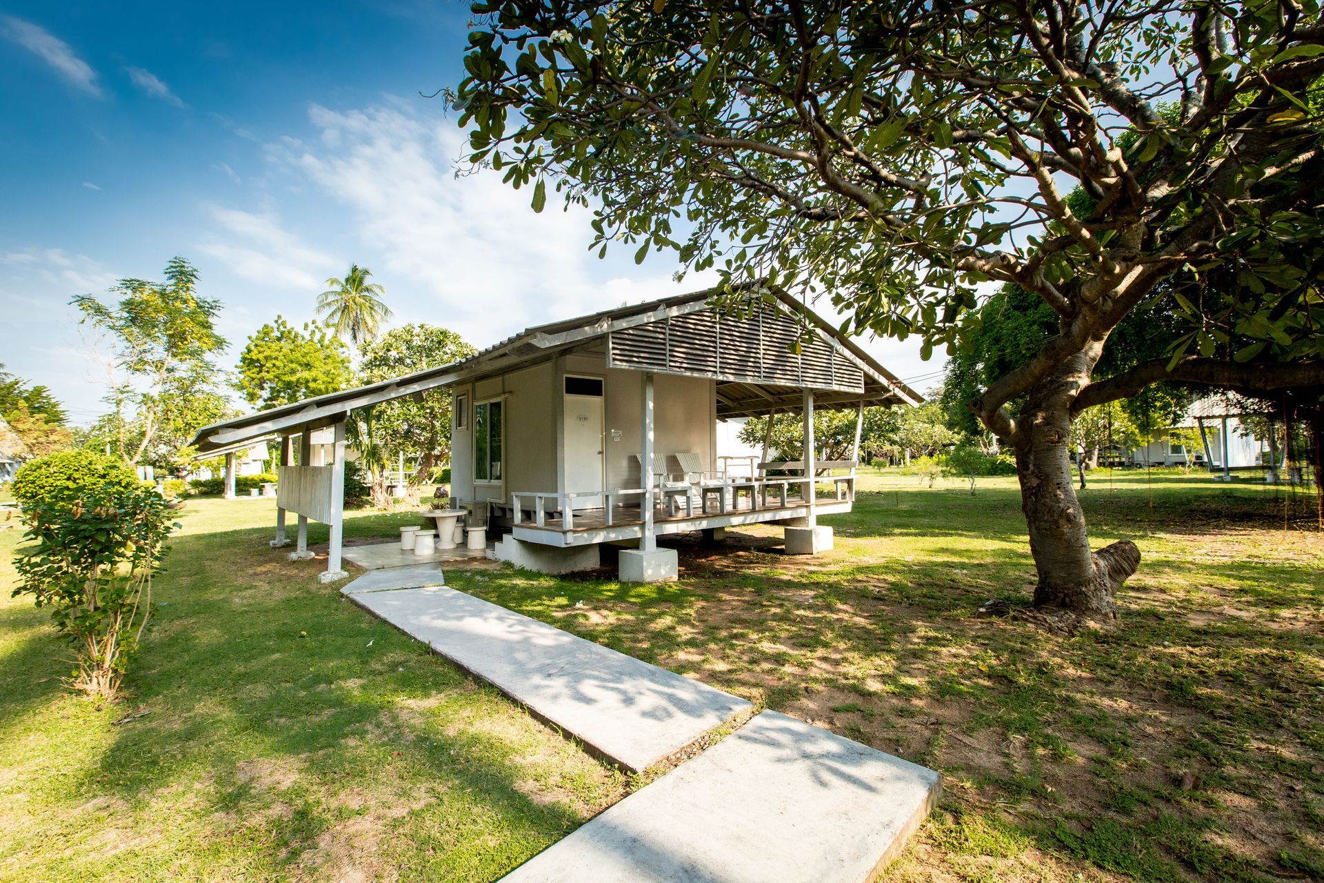 一座白色小别墅，带顶棚的门廊，坐落在绿草茵茵的草坪上。一条水泥小路通往别墅，头顶绿树成荫。
