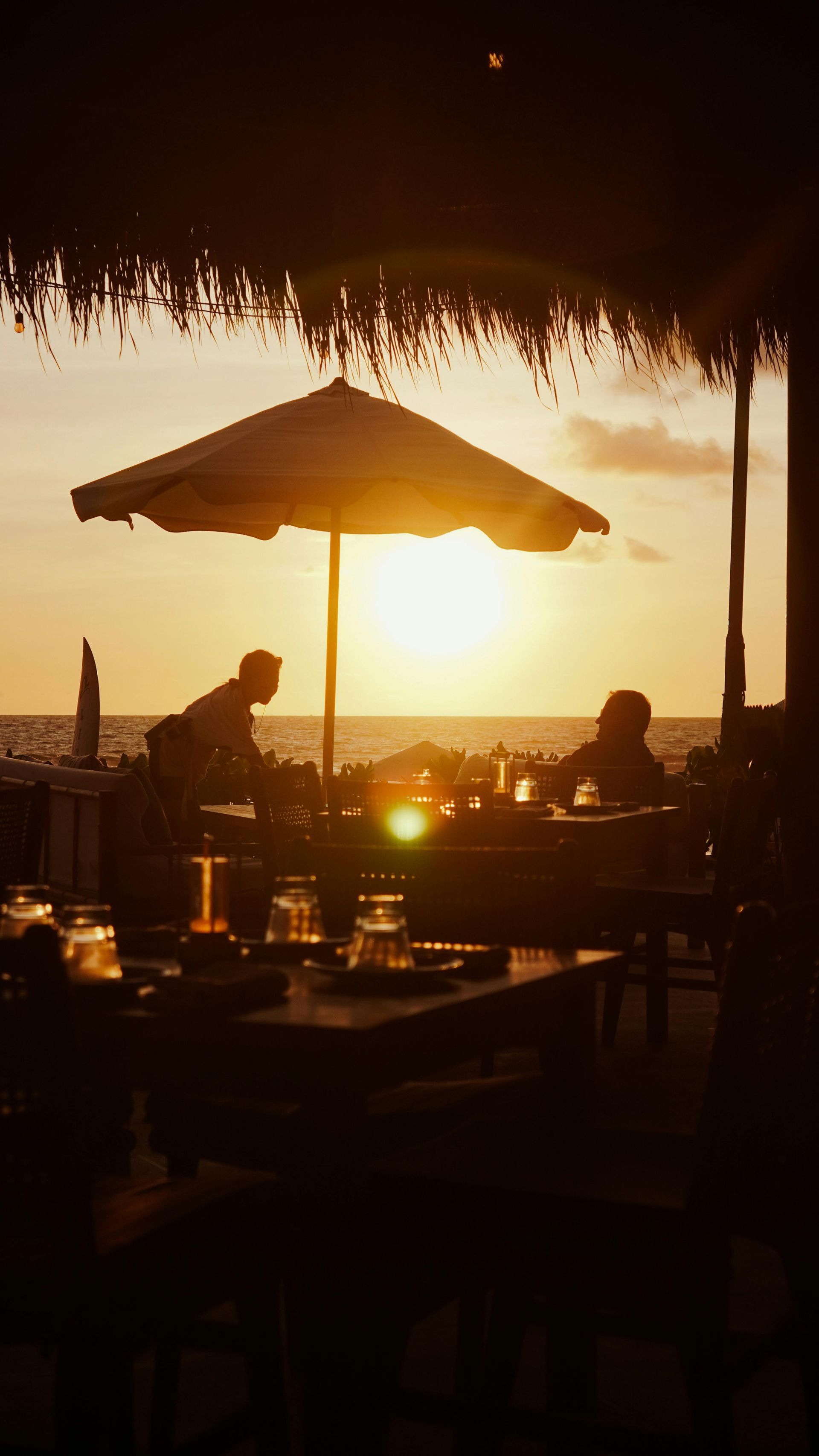 Restaurant tables silhouetted against a sunset. People dine under a large umbrella; a server works.