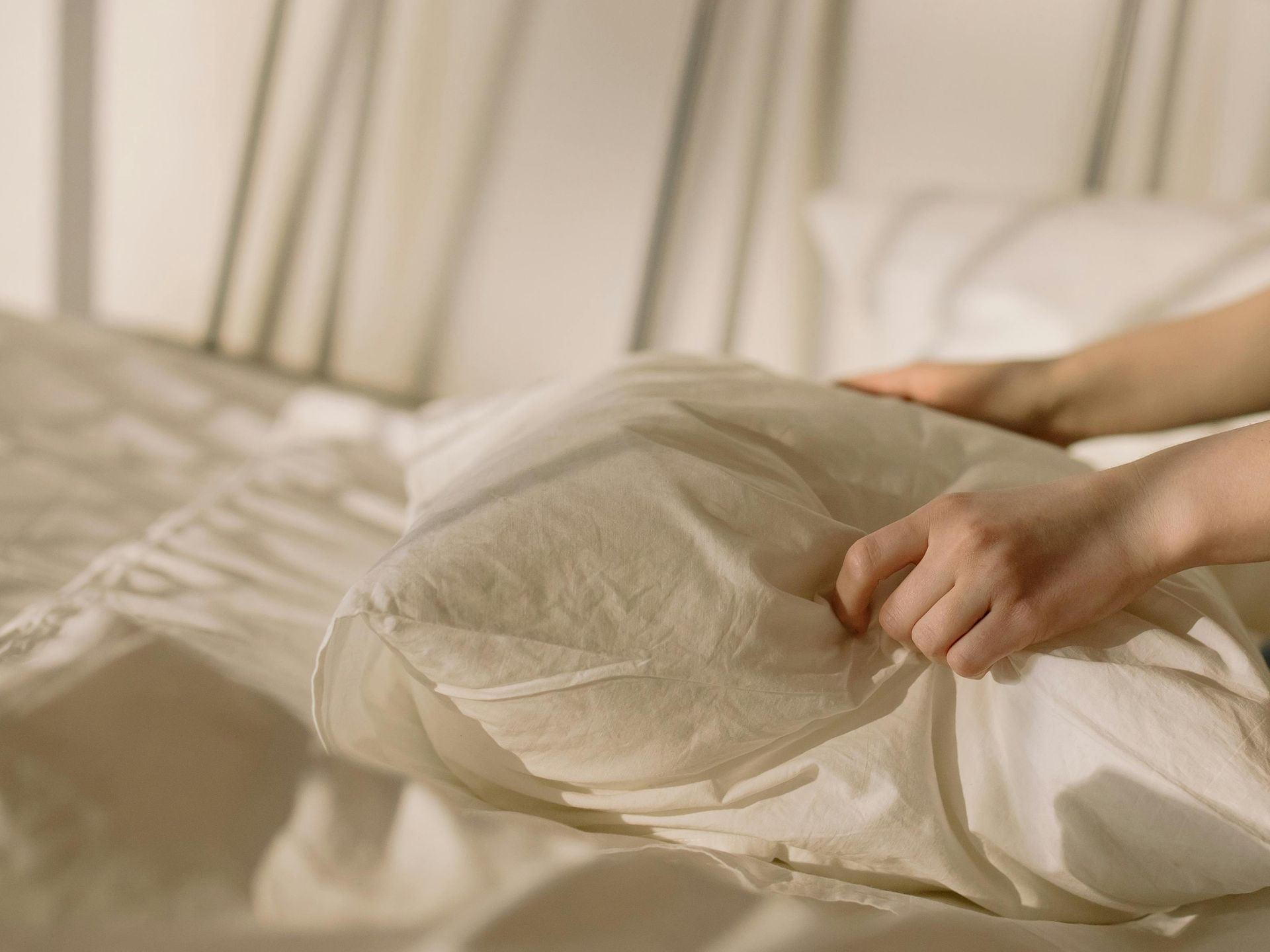 Hands arranging a pillow on a bed with white sheets. The background has a striped wall.