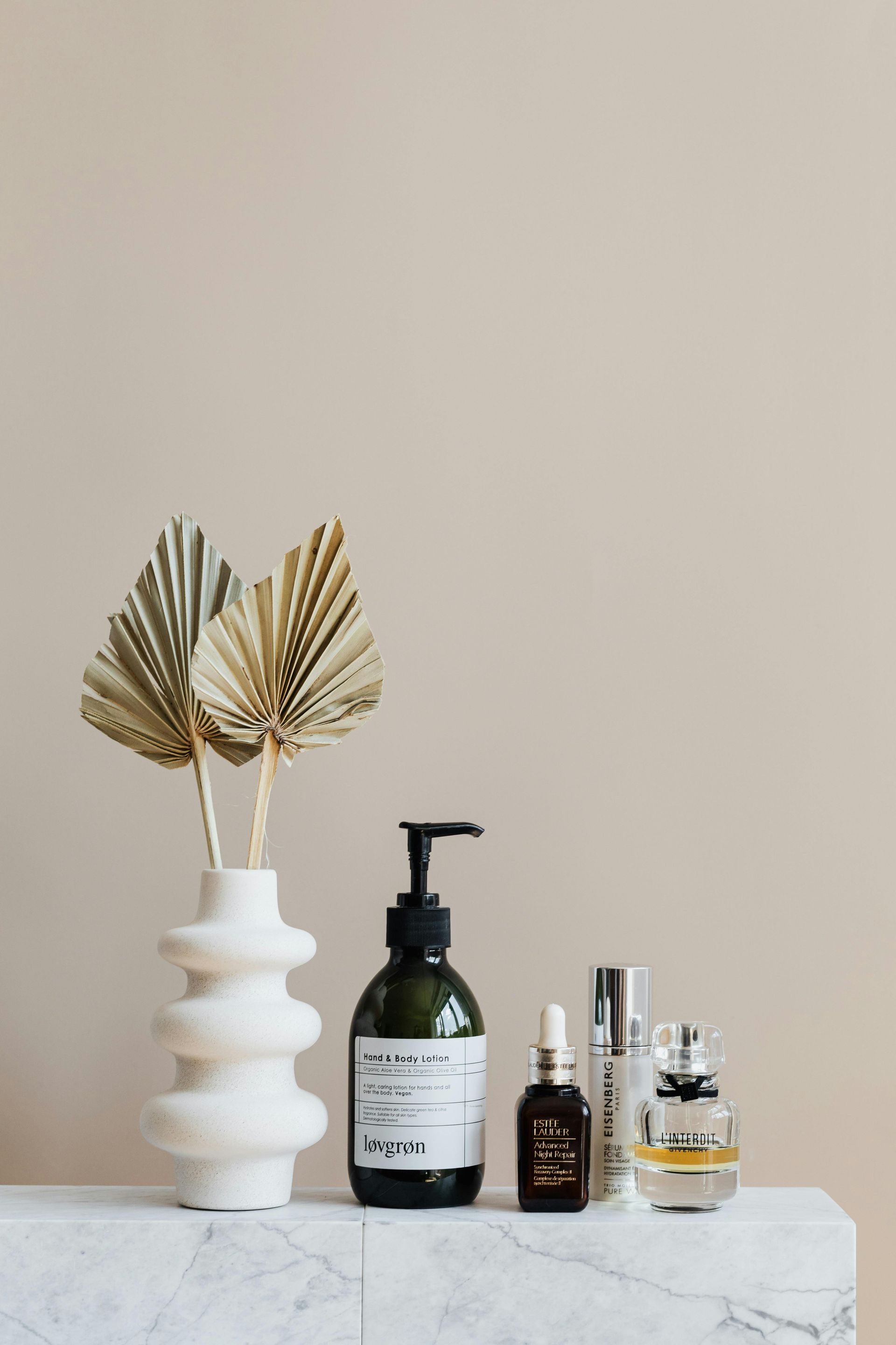Dried palm leaves in a white vase, skincare bottles on a marble surface, against a beige wall.