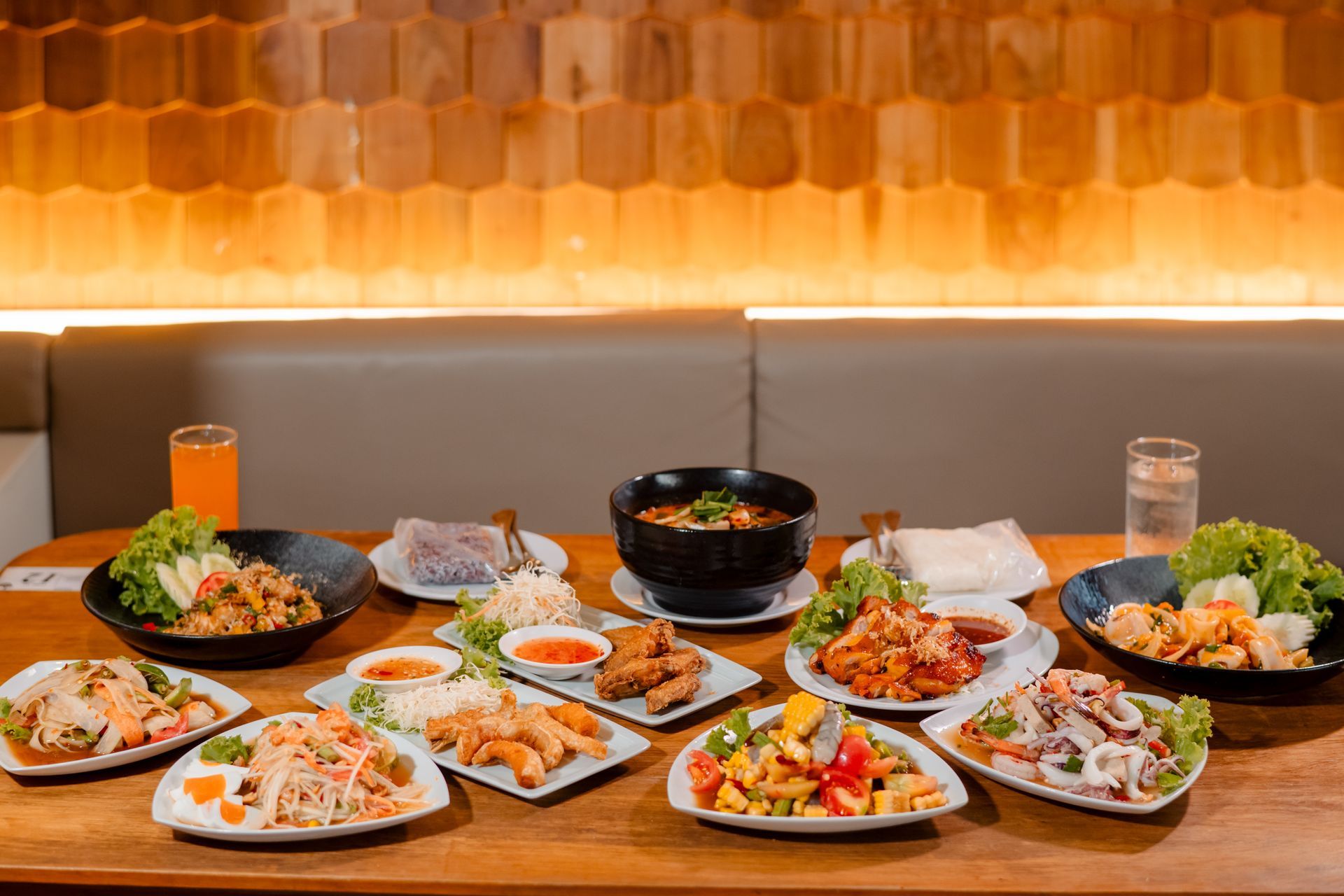 Wooden table with various Thai dishes, drinks, and a honeycombed wall in the background.