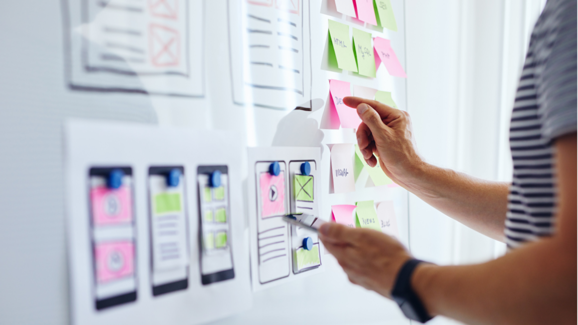 Person arranging sticky notes on a board with sketched website layouts, brainstorming ideas.
