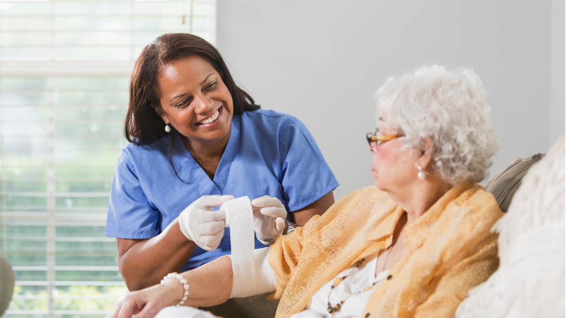A nurse in blue scrubs bandages an elderly woman's arm indoors. Both women are smiling.