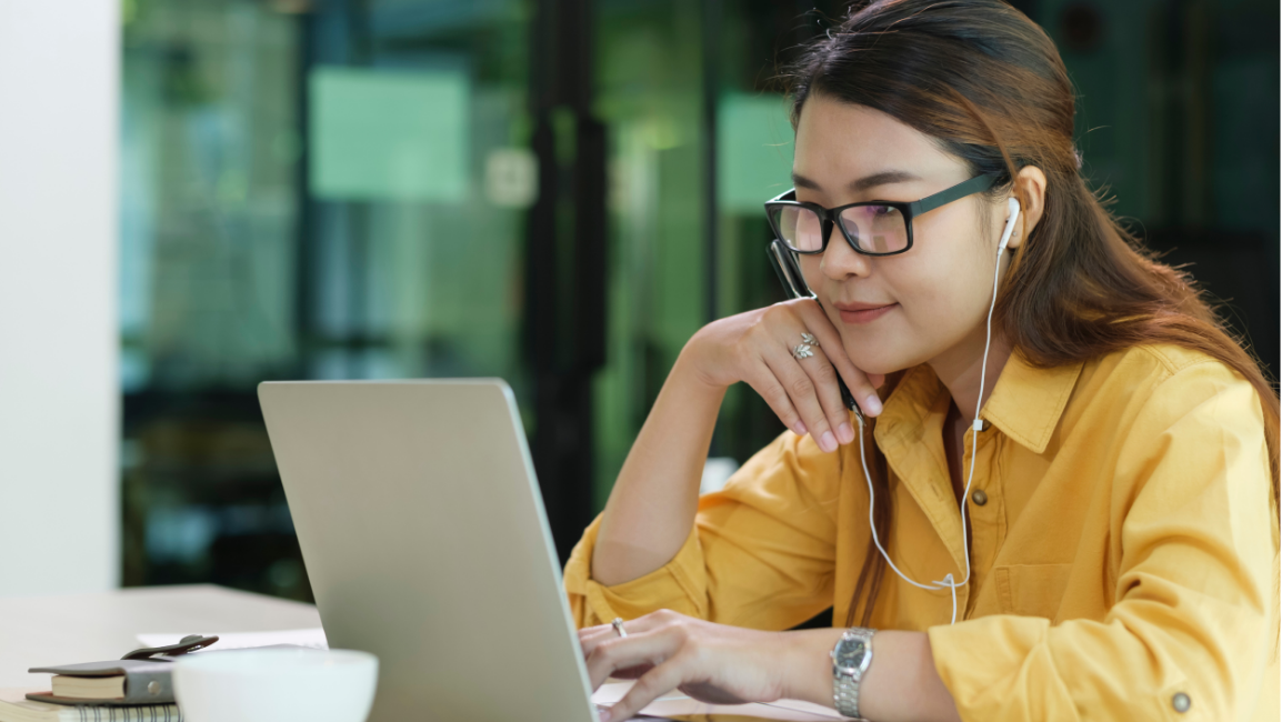 A woman with glasses and a yellow shirt is working on a laptop, wearing earbuds in a bright office.