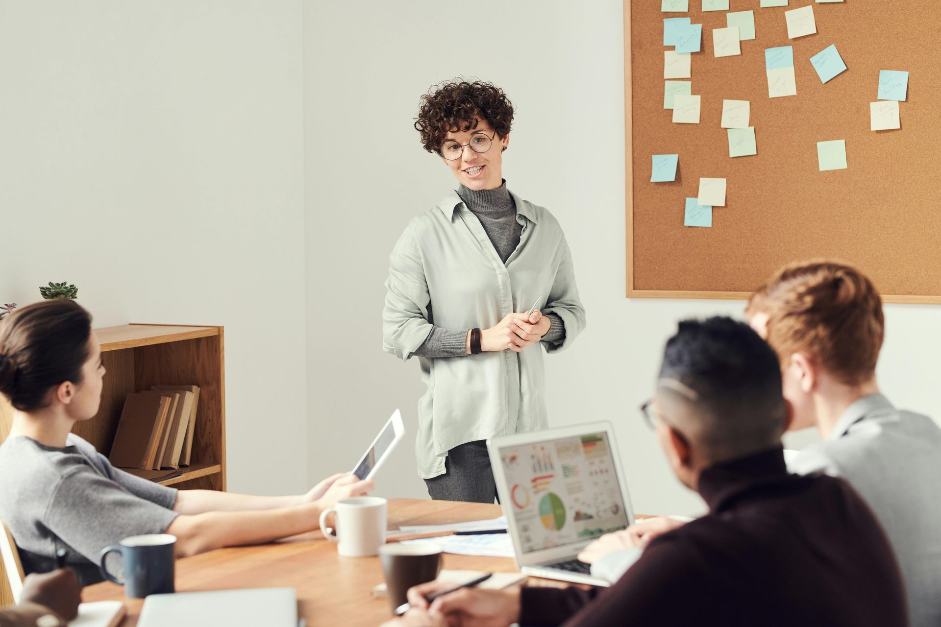 Woman presenting to colleagues in a meeting room, smiling, with laptops and sticky notes.