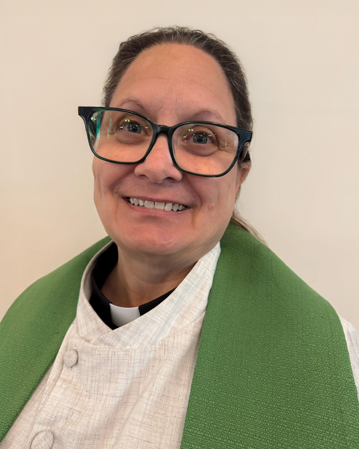 Woman in glasses and clerical collar, smiling, green stole against white wall.