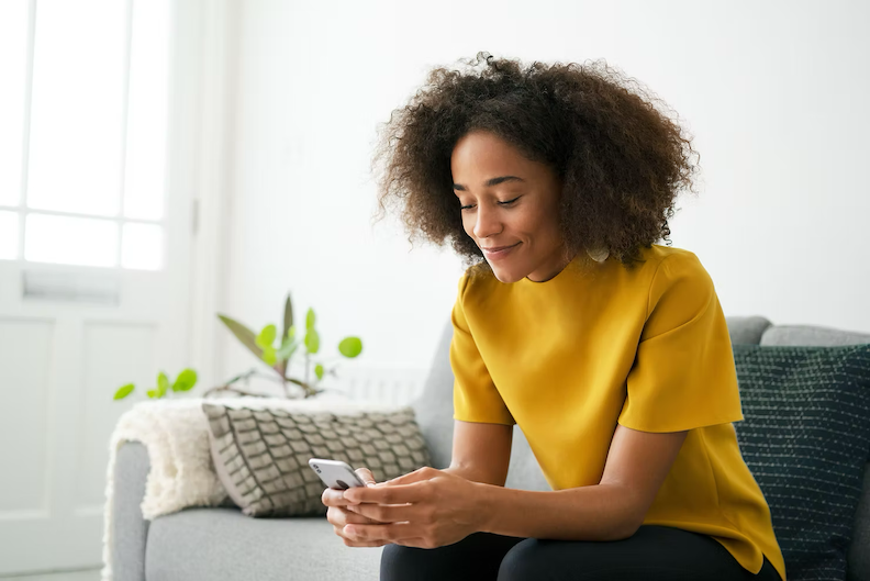Brown-skinned woman relaxing on a sofa, smiling quietly as she reads a supportive message on her pho