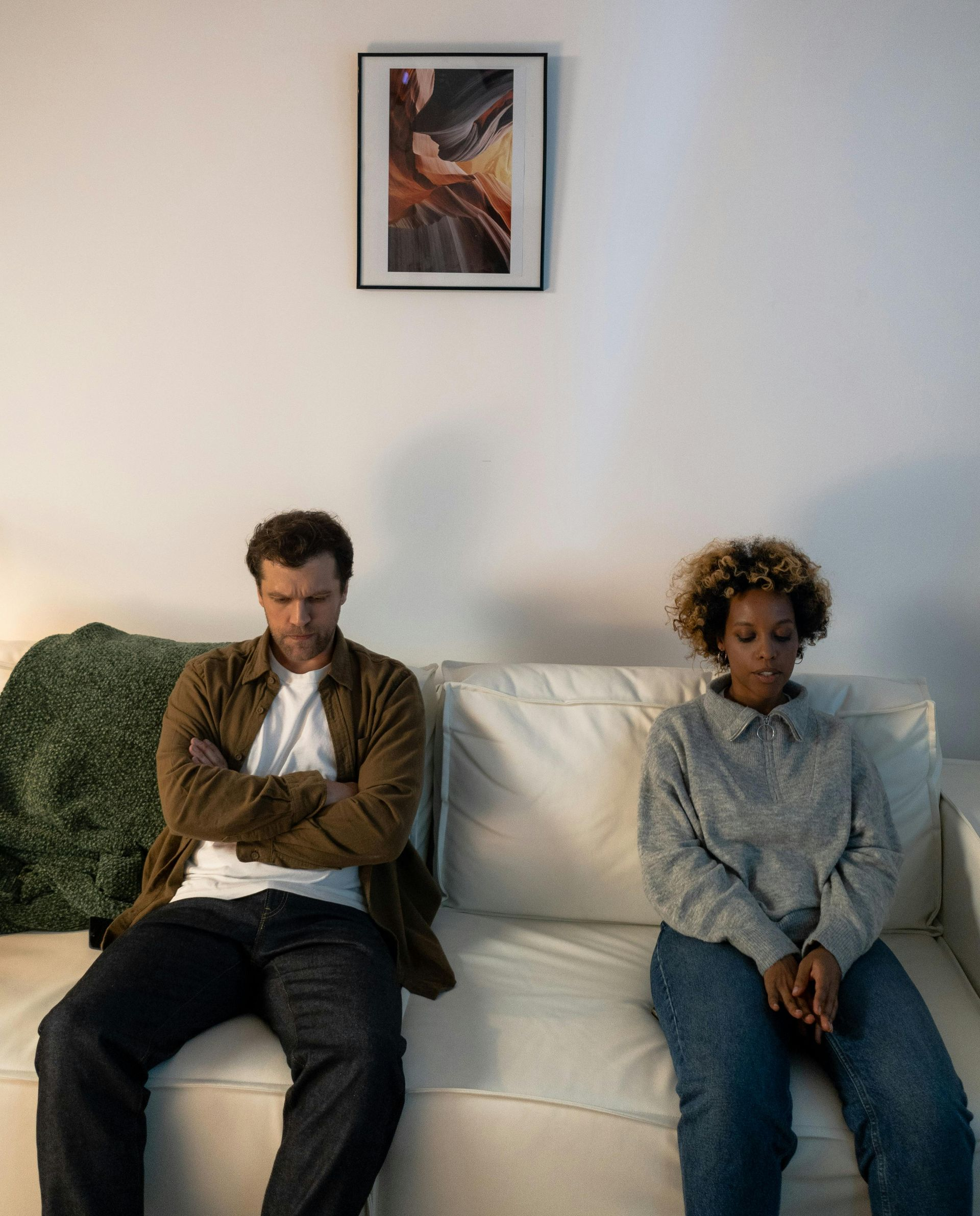 A couple sitting apart on a couch, both looking distant and upset, suggesting conflict or disconnect