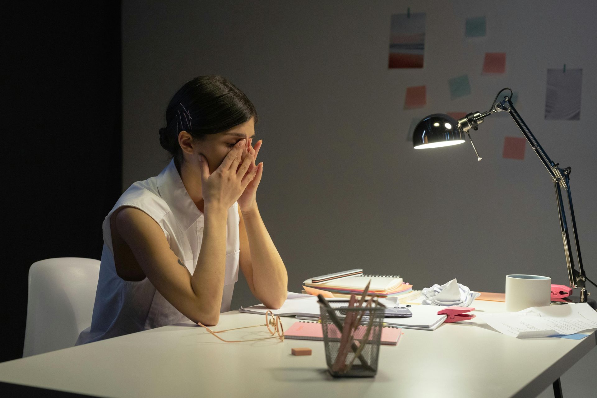 Woman of color sitting at a work space in  in a fatigued posture with her head bowed in her hands.
