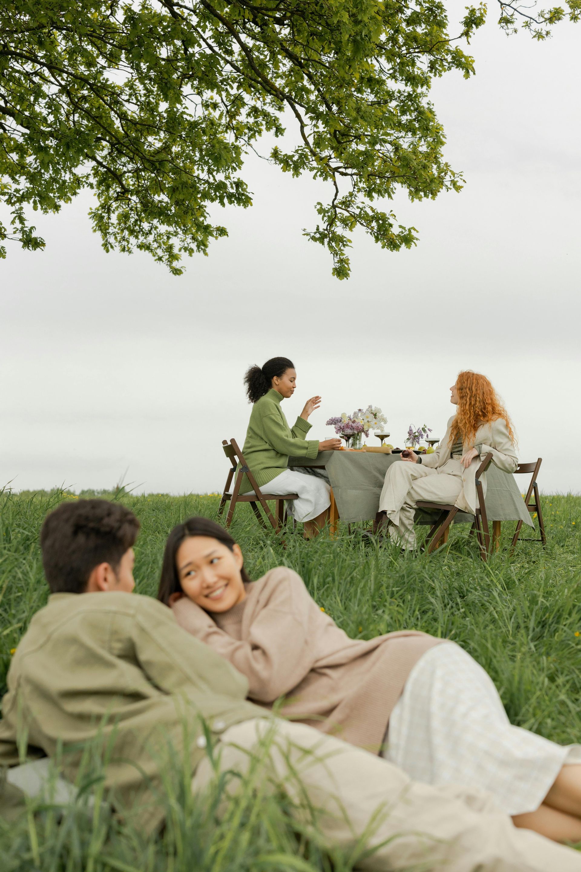 Two women sit at a small outdoor table with drinks and flowers, chatting in a grassy field, while two others relax nearby.