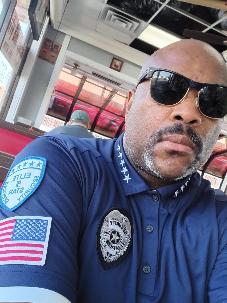 A Black man in a blue uniform with sunglasses, an American flag patch, and a badge, indoors.
