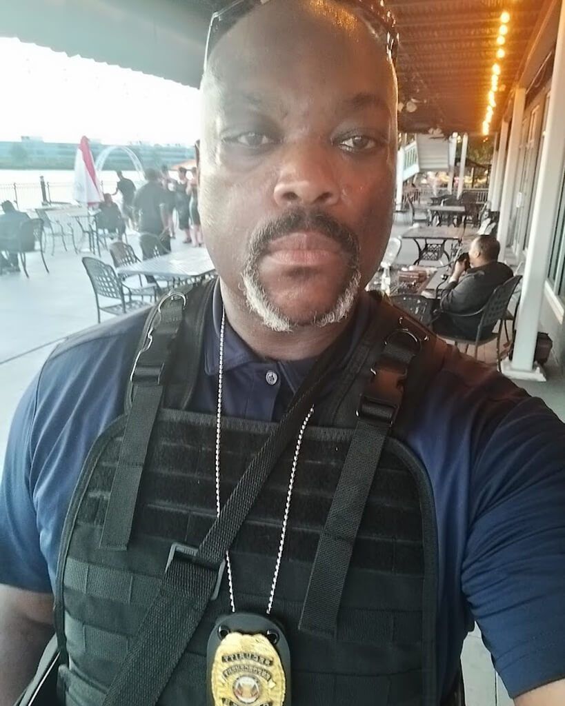 Man in blue shirt and vest, badge visible. Outdoors, blurred background.