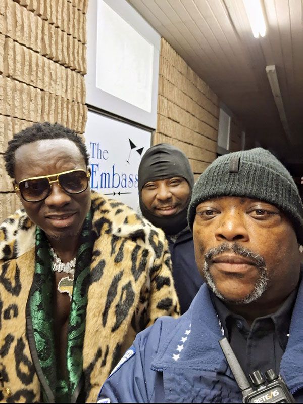 Three men pose for a selfie outside "The Embassy." Man in leopard coat, sunglasses; others in winter hats.