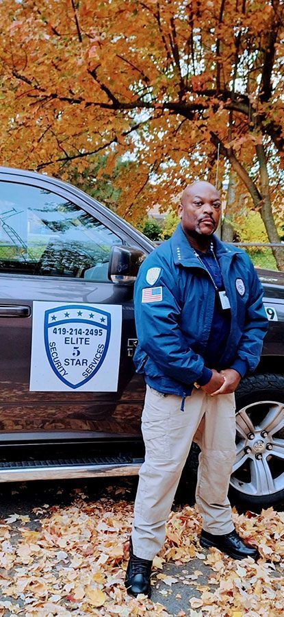 A man in blue jacket and tan pants stands by a car with fall foliage in the background.