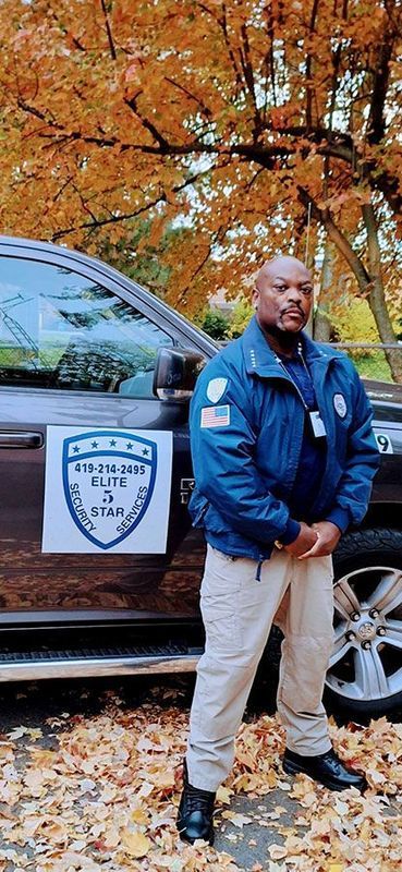 Man in blue jacket and tan pants stands beside a vehicle in autumn.