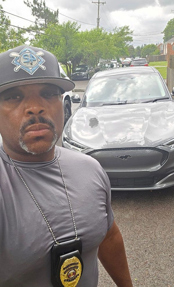 Man in hat and badge poses with a gray car on a street.