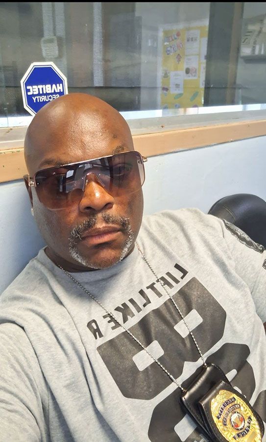 A bald Black man in sunglasses wearing a gray shirt with 