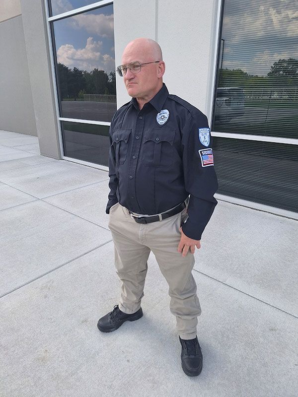Bald man in security uniform standing outside, hands in pockets.