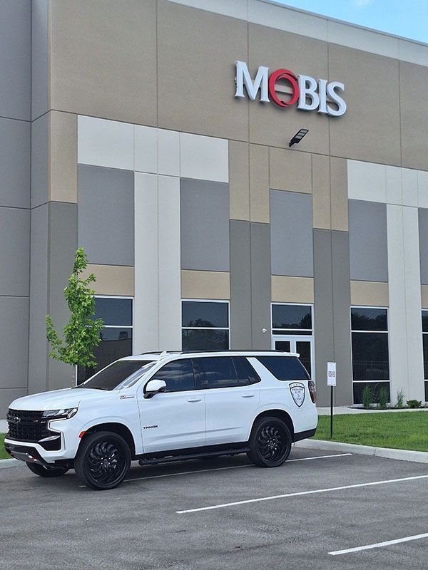 White SUV parked in front of MOBIS building. Gray and beige building exterior. Blue sky.