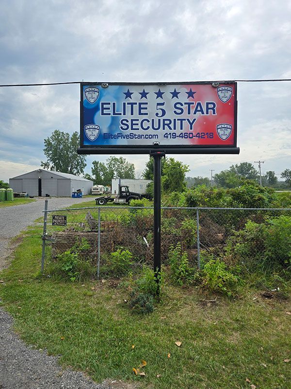 Sign for Elite 5 Star Security, blue and red, stars, phone number visible, in front of a building and fence.