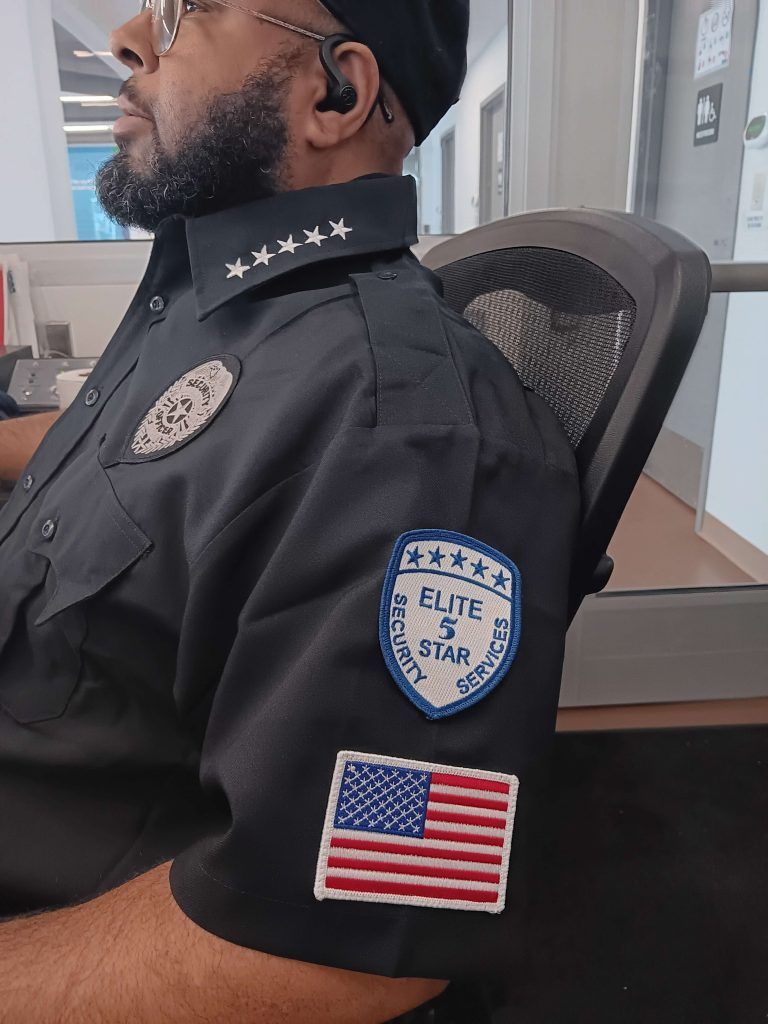 Security guard in black uniform with badges, US flag patch, and Elite 5 Star Service patch.