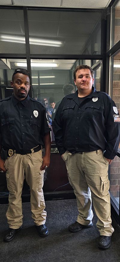 Two uniformed security guards standing inside a building. They are wearing dark blue shirts and khaki pants.